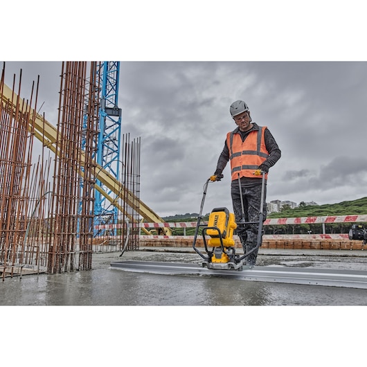 Worker uses DEWALT tool to smooth concrete at construction site, wearing safety gear and high-visibility vest.