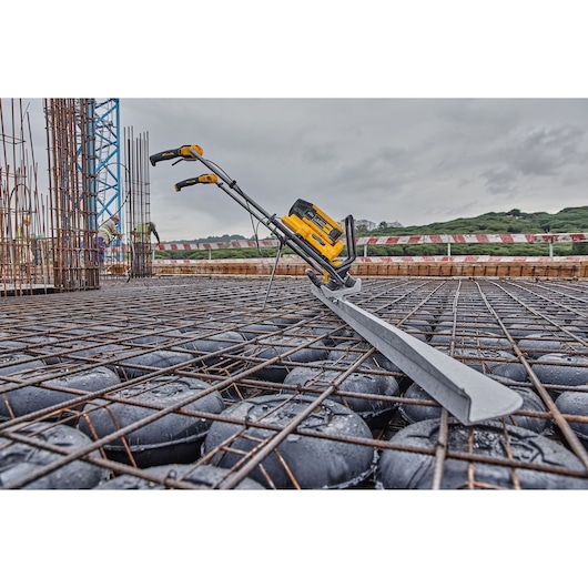 A DEWALT battery-powered concrete screed tool is placed on a construction site, positioned over a steel reinforcement grid and black plastic void formers. The tool is designed for smoothing and leveling concrete surfaces.