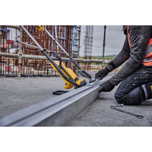 Worker uses DEWALT tool on metal beam at construction site, wearing gloves and safety vest.