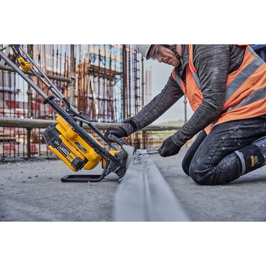 Worker uses yellow DEWALT tool to saw metal at construction site, wearing safety gear and orange vest.