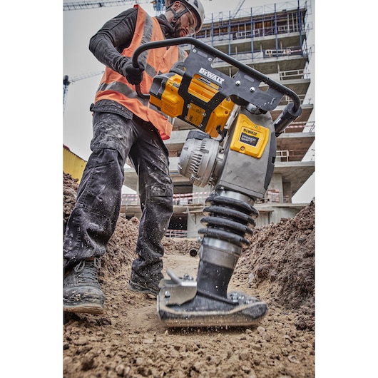 Worker uses DEWALT compactor DCPS660N on soil at construction site with buildings and scaffolding in the background.