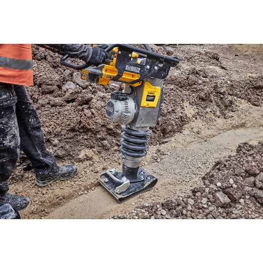 DEWALT rammer compacts soil on a construction site, operated by a worker in safety gear.