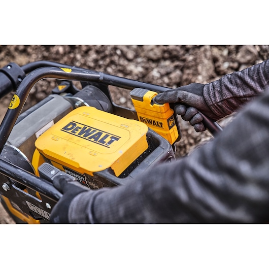 Close-up of gloved hands operating a yellow DEWALT tool outdoors, showing controls and branding.