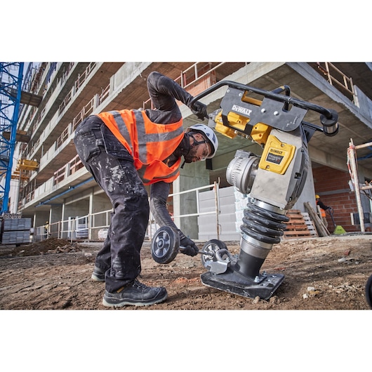 Worker uses a DEWALT compactor machine on a construction site, wearing safety gear and adjusting wheels.