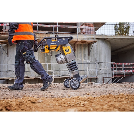 Worker walks with a DEWALT DCPS660N power tool on wheels at a construction site with dirt and concrete structures.
