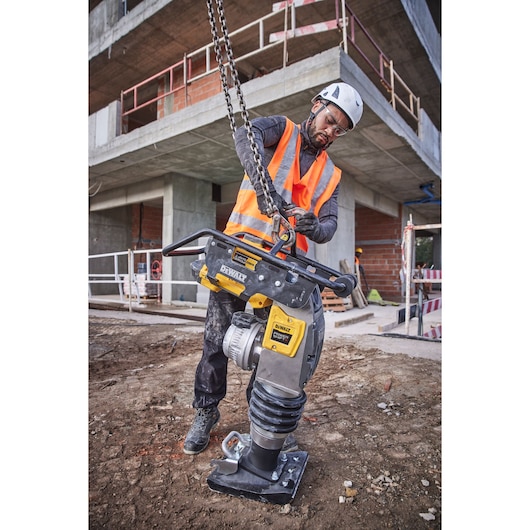Worker lifts DEWALT DCPS660N tool with chain at a construction site, wearing helmet and safety vest.