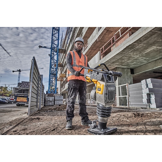 Person in safety gear using a DEWALT compactor at a construction site with cranes and a building in the background.