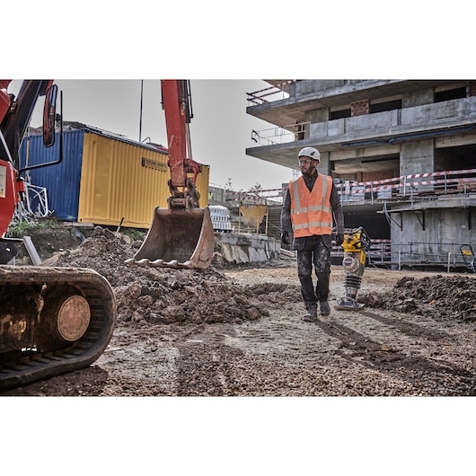 Worker in safety vest and helmet at construction site near excavator and DEWALT machinery.