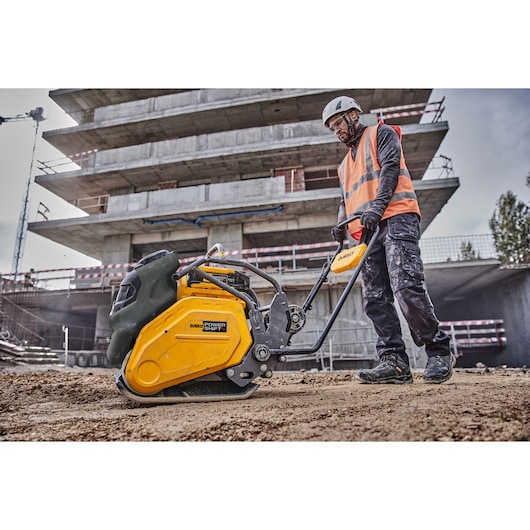 Worker uses DEWALT plate compactor at construction site, wearing safety gear and vest.