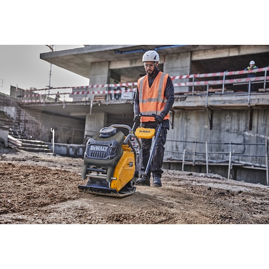 Worker uses a DEWALT plate compactor at a construction site, wearing safety gear and orange vest.