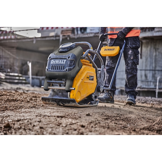 DEWALT plate compactor used on dirt by a person at a construction site, close-up of machinery and worker's legs.