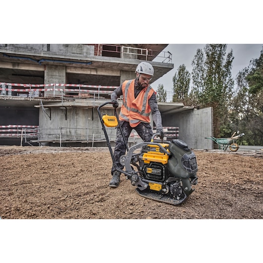 Worker with DEWALT plate compactor DCPS7154N on construction site, wearing safety gear and orange vest.