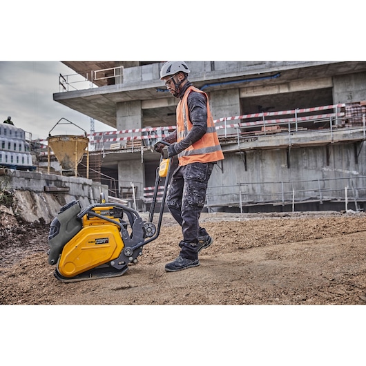 Worker uses DEWALT compactor DCPS7154N on soil at a construction site, wearing safety gear and high-visibility vest.