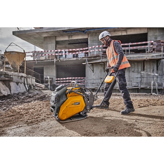 Worker uses a DEWALT plate compactor on a construction site with unfinished buildings in the background.