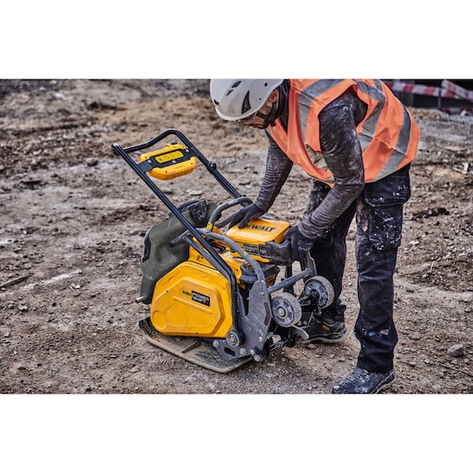 Worker in safety gear operates a yellow DEWALT plate compactor on a construction site with dirt ground.