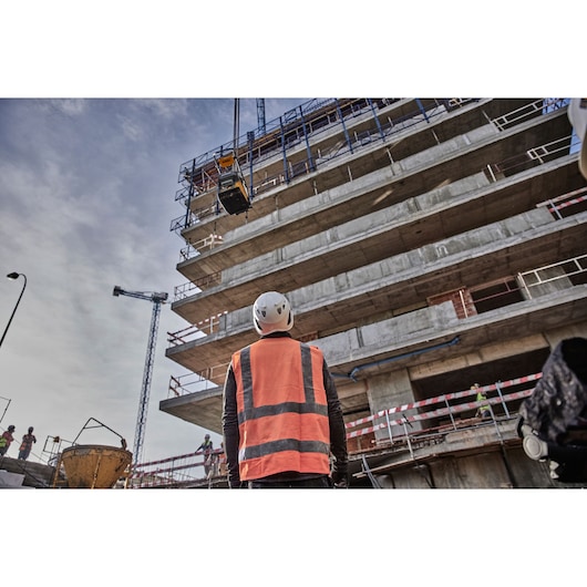 Worker in orange vest and helmet at a construction site, looking up at a building with crane overhead.