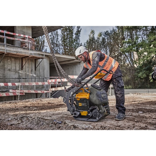 Worker with helmet and vest operates DEWALT compactor on a construction site with safety barriers.