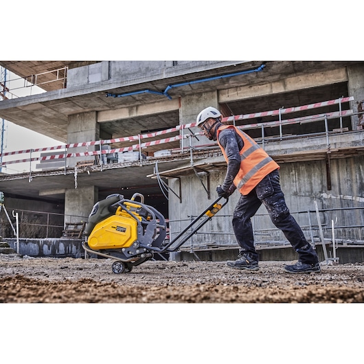 Worker uses a DEWALT compacting tool on soil at a construction site, wearing safety gear and a high-visibility vest.