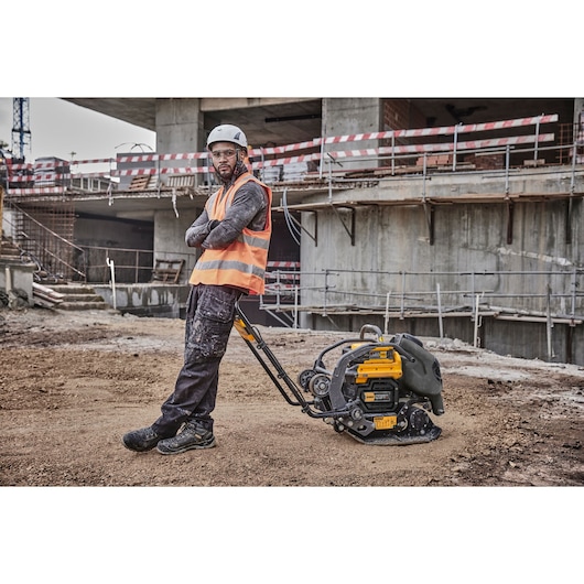 Worker in safety gear using a DEWALT compactor DCPS7154N at a construction site with unfinished building.