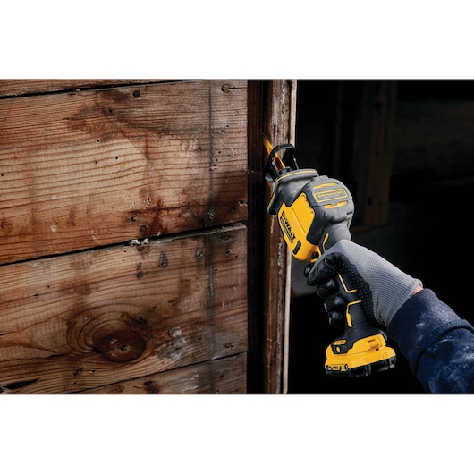 Gloved hand using a DEWALT cordless reciprocating saw on a wooden surface indoors.