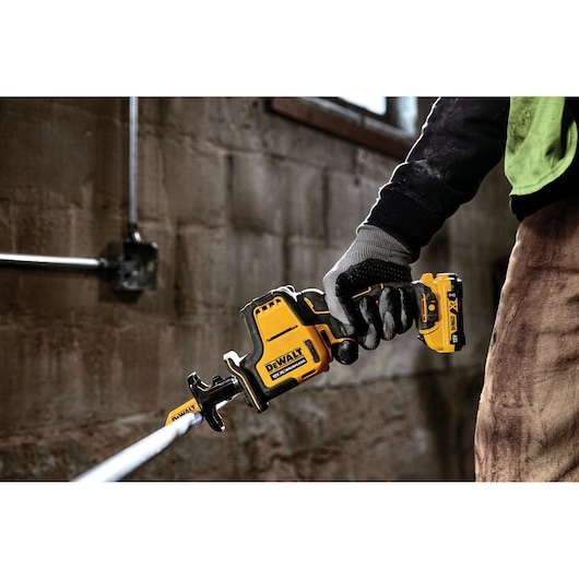 Person using a DEWALT DCS312N cordless reciprocating saw to cut a pipe in a workshop.