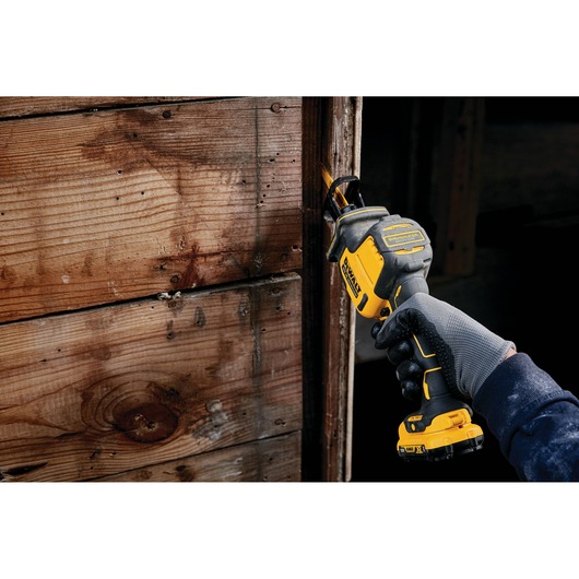Person uses a DEWALT reciprocating saw to cut wood near a wall. Gloved hand holds the yellow and black tool.