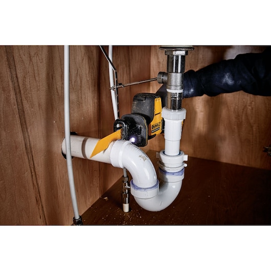 DEWALT tool cutting white PVC plumbing pipe under a sink in a wooden cabinet.