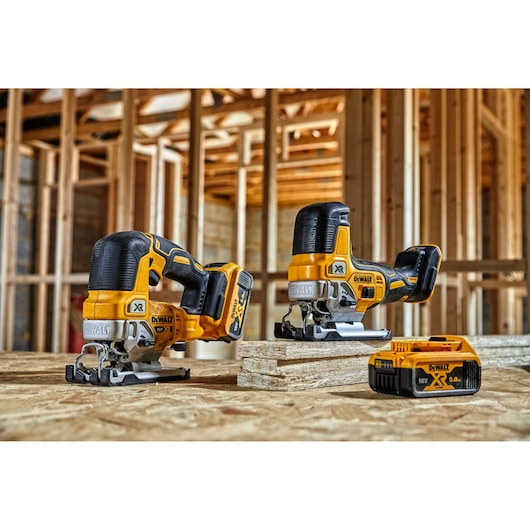 Two DEWALT cordless jigsaws and a battery on a wooden surface at a construction site with wooden framing.