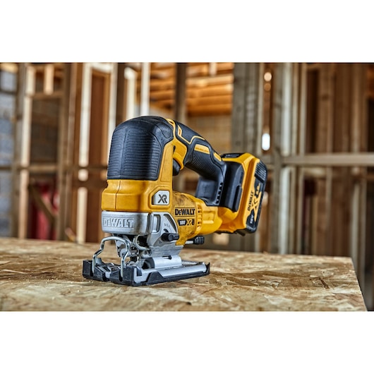 DEWALT cordless jigsaw placed on plywood in a construction site with wood framing visible in the background.