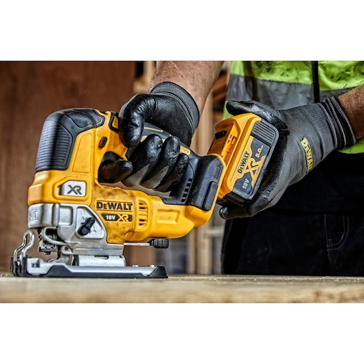 A gloved hand holds a DEWALT DCS334P2 18V cordless jigsaw on a worktable in a workshop setting.