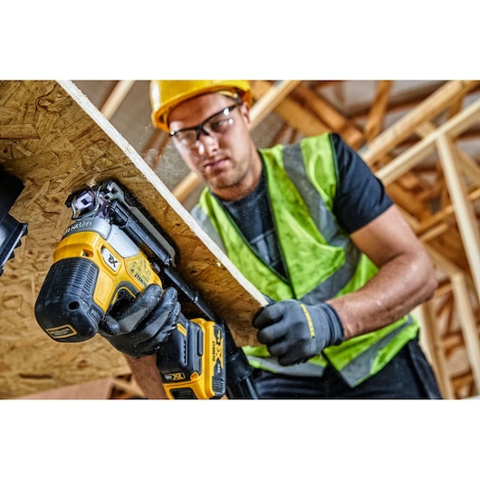 Worker uses a DEWALT cordless jigsaw to cut wood panel at a construction site, wearing safety gear.