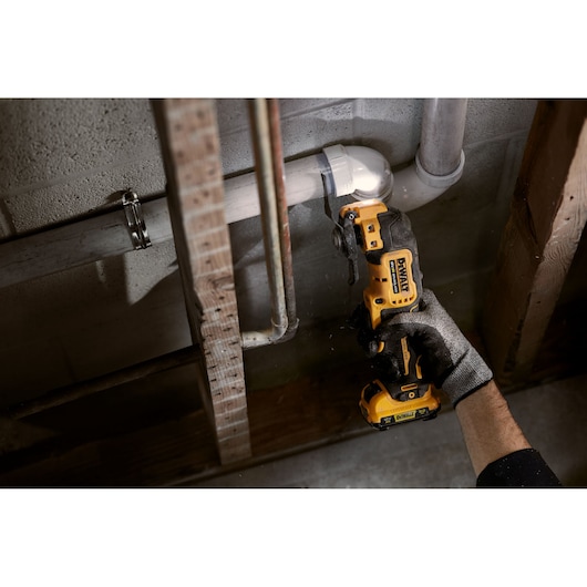 Person uses a DEWALT cordless oscillating tool to cut a pipe against a concrete wall in a construction setting.