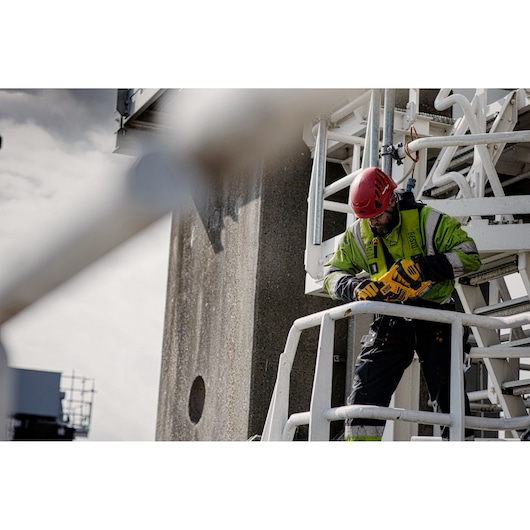 A construction worker wearing a safety helmet and high-visibility jacket is using a DEWALT power tool while standing on an industrial metal platform. The worker is secured with a safety harness.