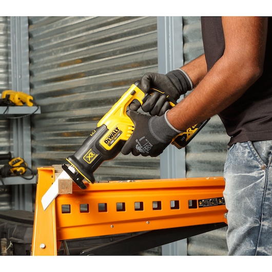 Person uses DEWALT DCS367P2 reciprocating saw to cut wood on an orange metal workbench in a workshop.