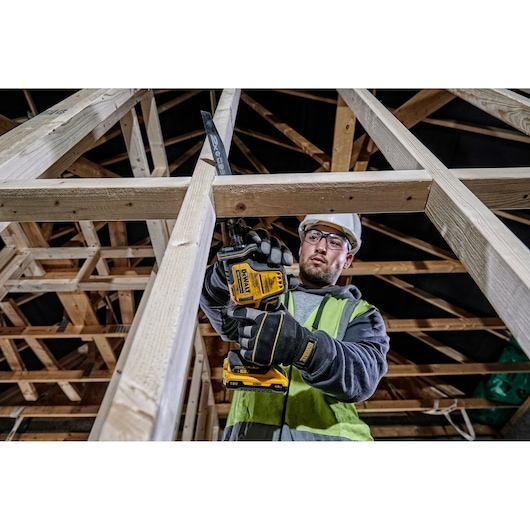 Worker uses a DEWALT cordless saw to cut wood framing at a construction site, wearing gloves and safety gear.