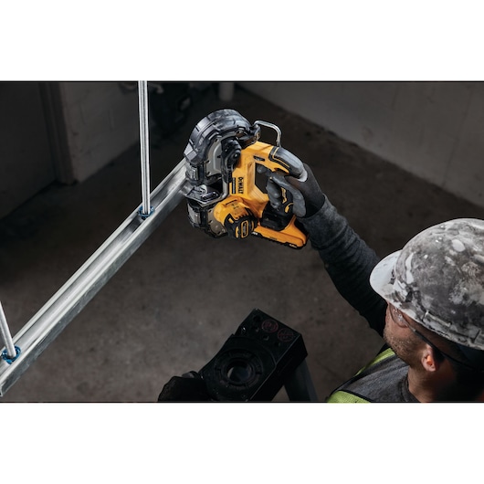 Worker uses yellow DEWALT cordless bandsaw to cut metal pipe in industrial setting.