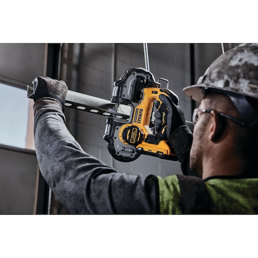 Person using a DEWALT cordless band saw to cut a metal pipe indoors, wearing gloves and a helmet.