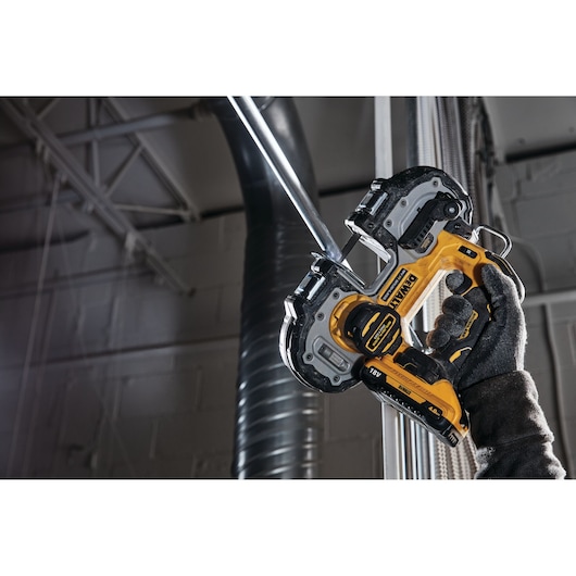 A DEWALT cordless band saw cutting a metal pipe, held by a gloved hand in an industrial setting.
