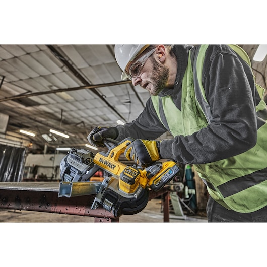 Person using DEWALT DCS378N band saw to cut metal in an industrial workshop, wearing safety gear.