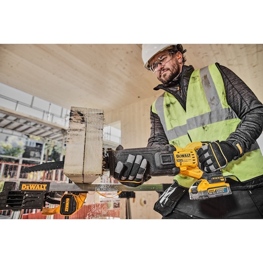 A construction worker wearing safety gear, including a helmet, gloves, and a high-visibility vest, is using a DEWALT cordless reciprocating saw (model DCS386H2T) to cut through a thick piece of wood on a building site.