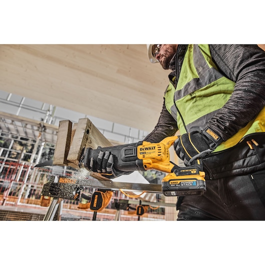 A construction worker using a DEWALT DCS386H2T cordless reciprocating saw to cut a piece of wood at a building site, wearing safety gear including gloves, vest, and helmet.