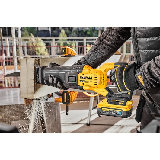 A person using a DEWALT cordless reciprocating saw to cut through a wooden beam, with sawdust flying. The saw features a DEWALT POWERSTACK battery and the scene takes place at a worksite.