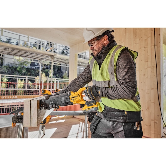 A person wearing a safety vest and hard hat is using a DEWALT cordless reciprocating saw (model DCS386H2T) to cut a piece of wood on a construction site.
