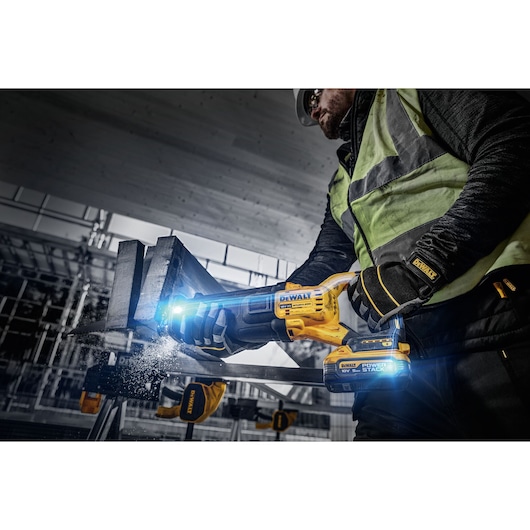 A worker using a DEWALT DCS386H2T cordless reciprocating saw to cut through a piece of metal at a construction site. The tool features a DEWALT POWERSTACK battery and is emitting bright light during operation.