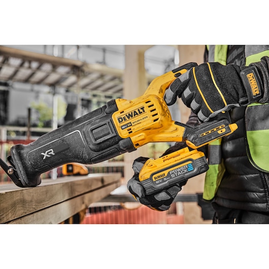 A close-up of a DEWALT XR 18V cordless reciprocating saw with a POWERSTACK battery, being held by a person wearing DEWALT-branded gloves and a safety vest on a construction site.