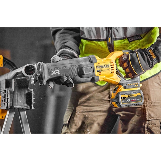 Worker using a DEWALT cordless reciprocating saw with battery to cut a metal pipe indoors.