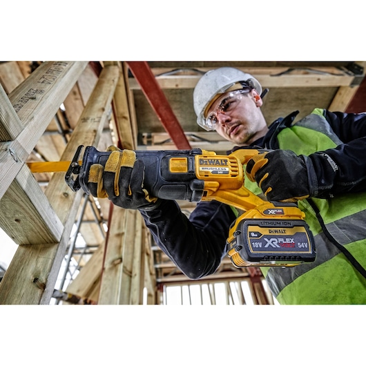 Worker uses DEWALT DCS389NT reciprocating saw to cut wood at construction site, wearing gloves and safety gear.