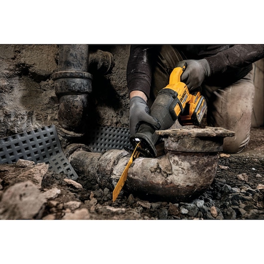 Person uses a DEWALT reciprocating saw to cut through a rusted pipe in a construction or repair setting.