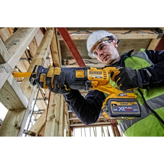 Worker using DEWALT DCS389T2 cordless reciprocating saw on wood frame at construction site.