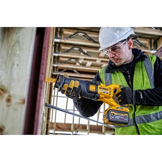 Worker using DEWALT DCS389T2 cordless saw at a construction site, wearing gloves, vest, and helmet.